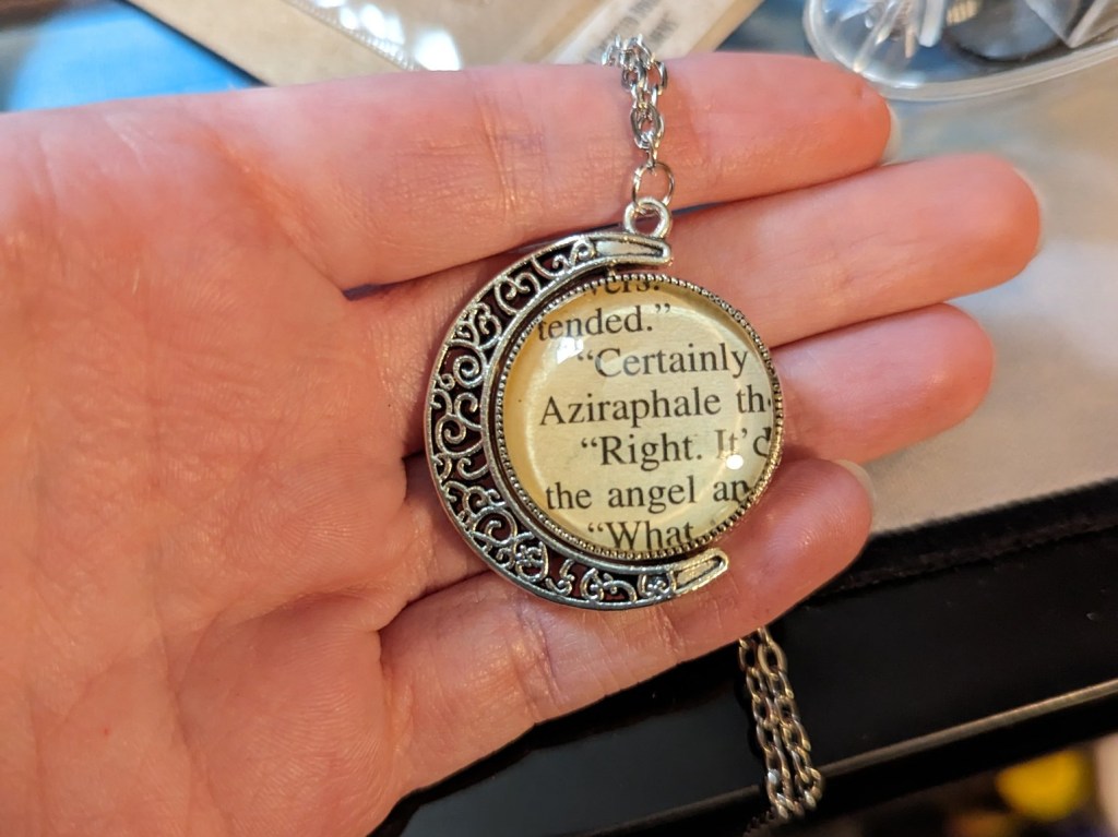 A silver necklace in the shape of a crescent moon. There is a clear sphere held in it. One side has text that includes "Aziraphale" and the other side "Crowley."