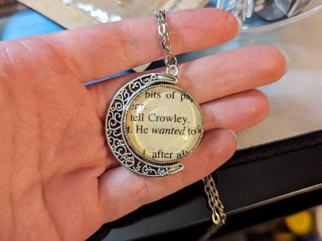 A silver necklace in the shape of a crescent moon. There is a clear sphere held in it. One side has text that includes "Aziraphale" and the other side "Crowley."