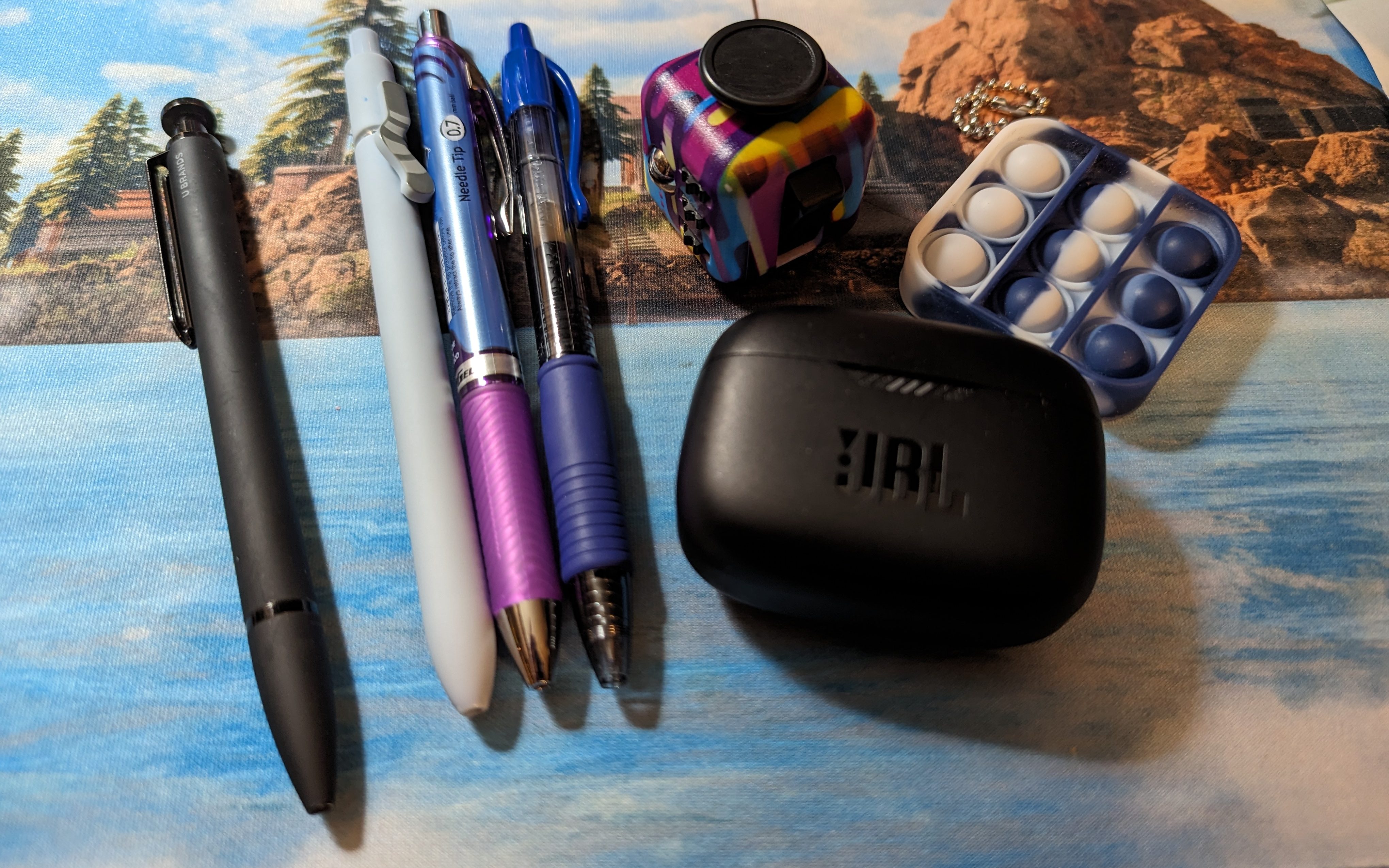 Two different fidgets, a wireless earbuds case, and 4 pens of different types. One of the pens has a little kitty tail on it.